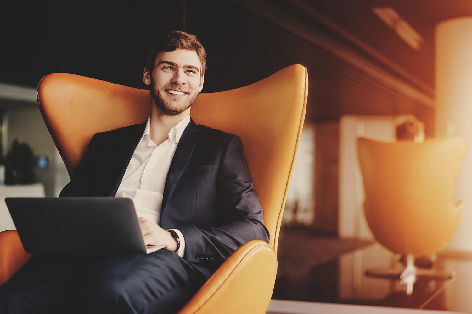 Молодой улыбающийся успешный человек предприниматель в формальном бизнес-люксе с бородкой сидит на оранжевом кресле с ноутбуком в роскошном офисном интерьере