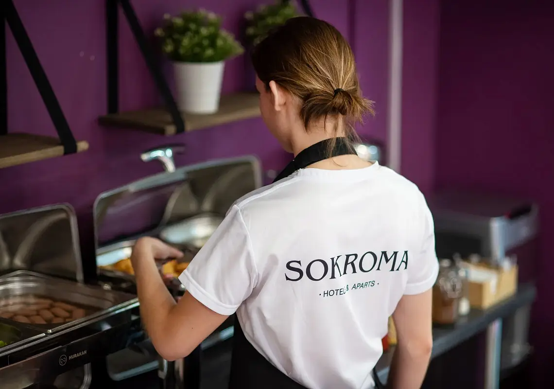 Сотрудница отеля в футболке с логотипом «Sokroma» пополняет блюда на шведском столе