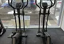 Два черных эллиптических тренажера с цифровыми панелями, установленные в спортзале с большими окнами